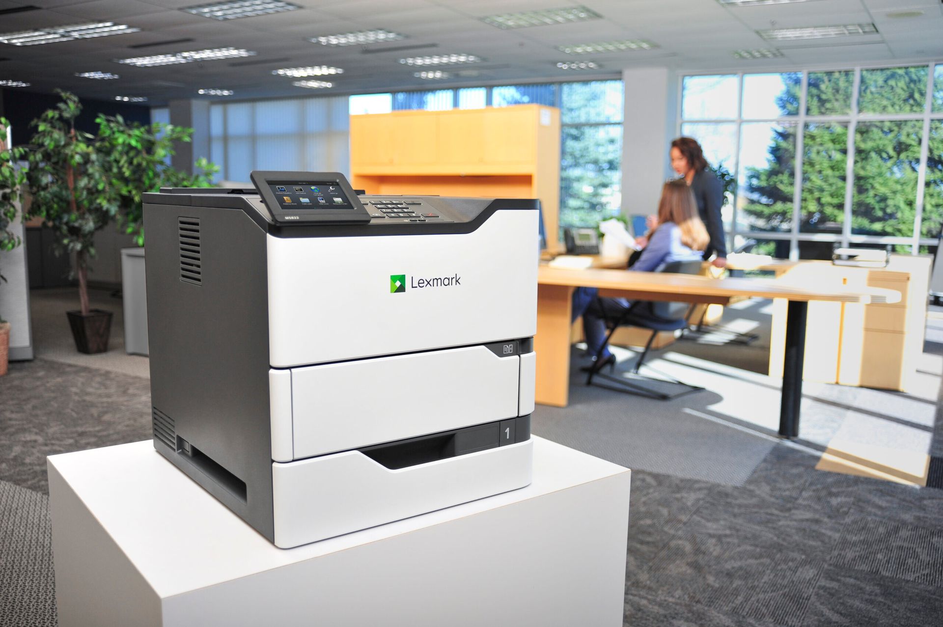 Large office printer on a white pedestal in a modern workspace, with a person sitting at a desk in the background.