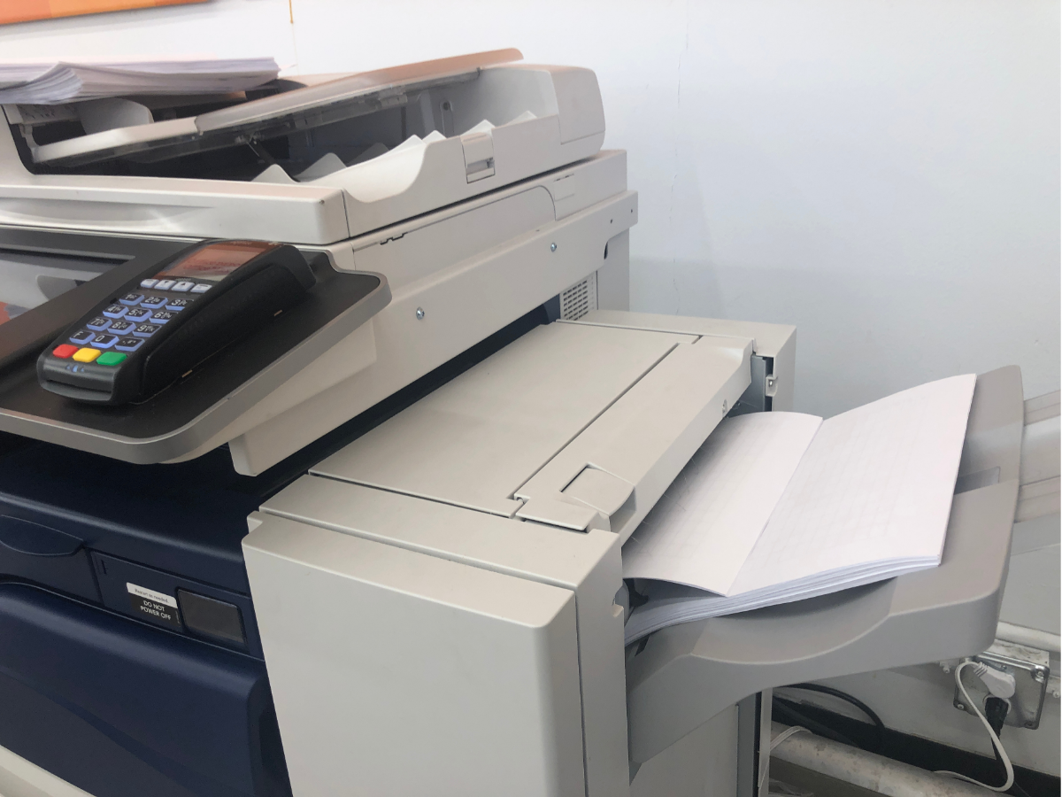 Office copier/printer with paper trays and a control panel, printing a stack of pages.