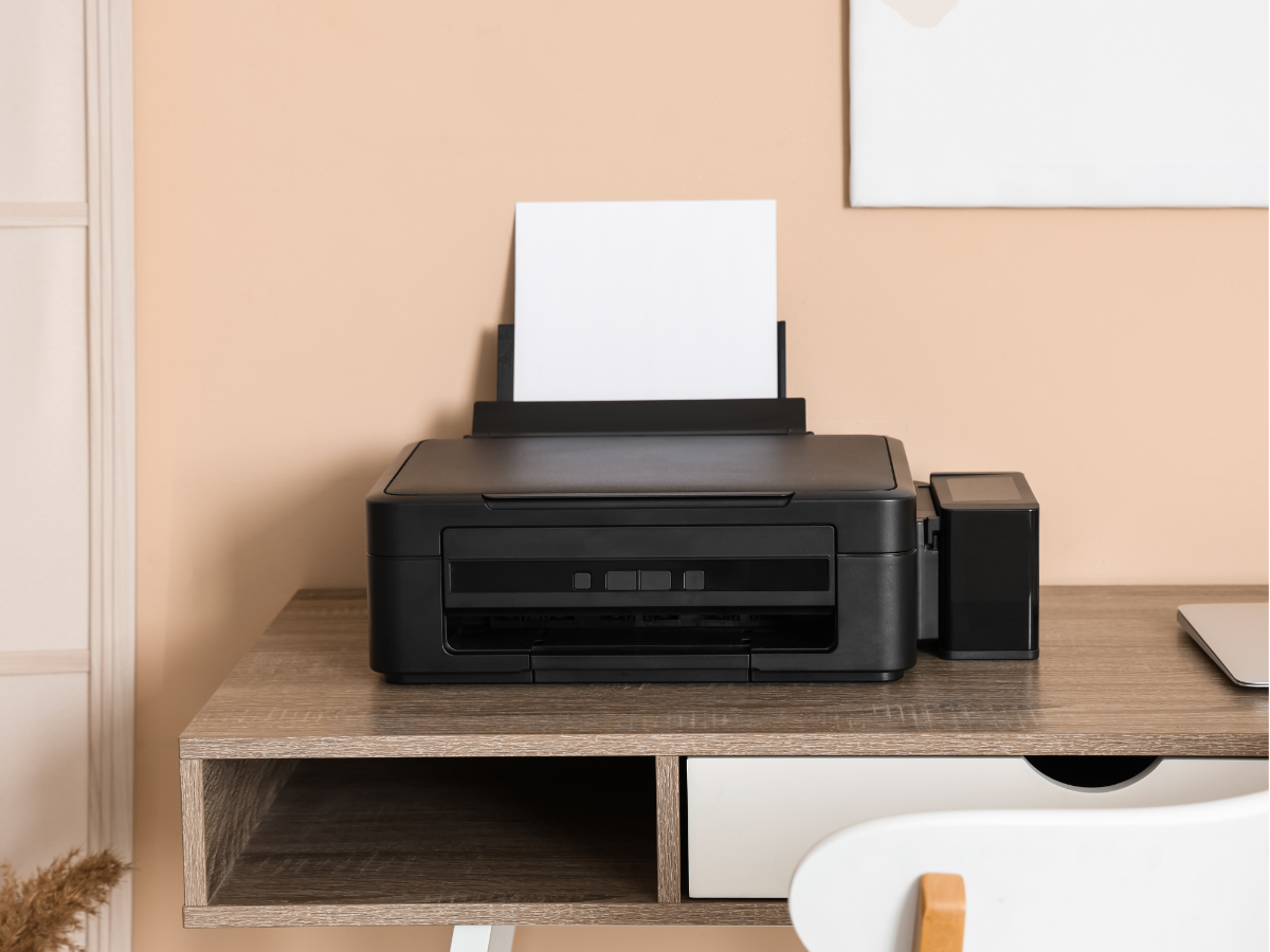 Black printer on a wooden desk with paper loaded, beside a small black box in a room with peach walls.