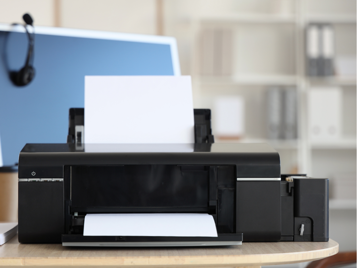 Black printer with paper loaded on a desk beside a blue laptop in an office setting