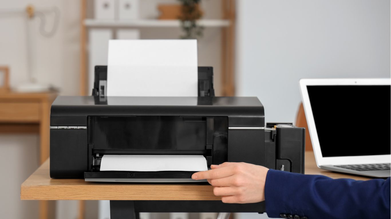 Black printer printing a page on a desk, with a hand reaching to remove the paper beside a laptop.