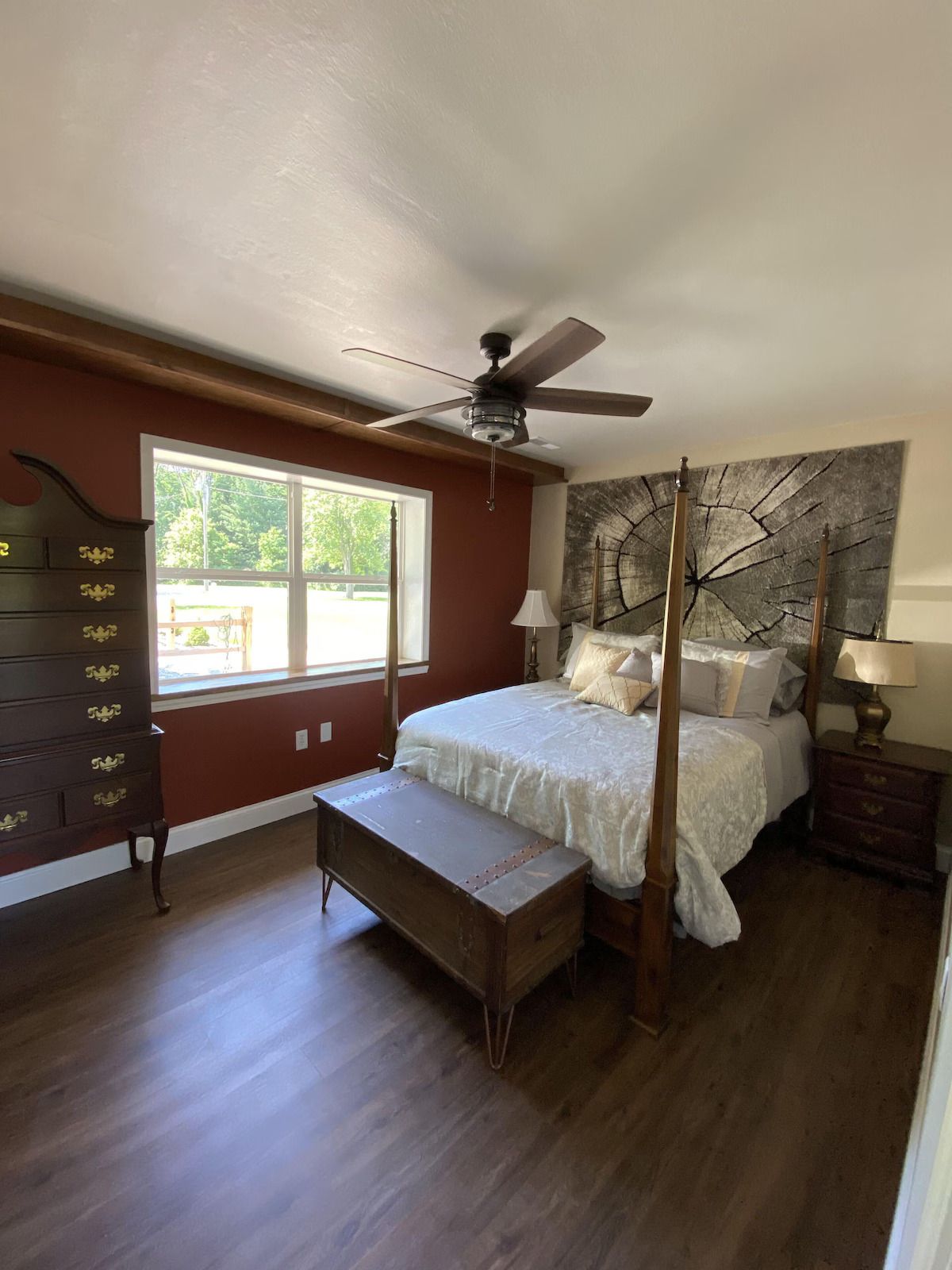 A bedroom with dark red walls, a four-poster bed, a large chest at the foot of the bed, and a dresser beside a window.