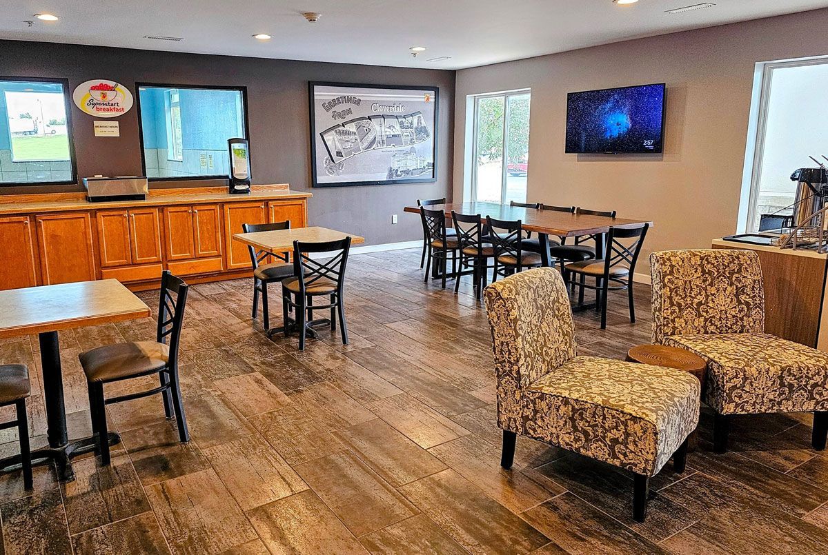 A hotel breakfast area with wooden cabinets, multiple small tables with chairs, and patterned accent chairs.