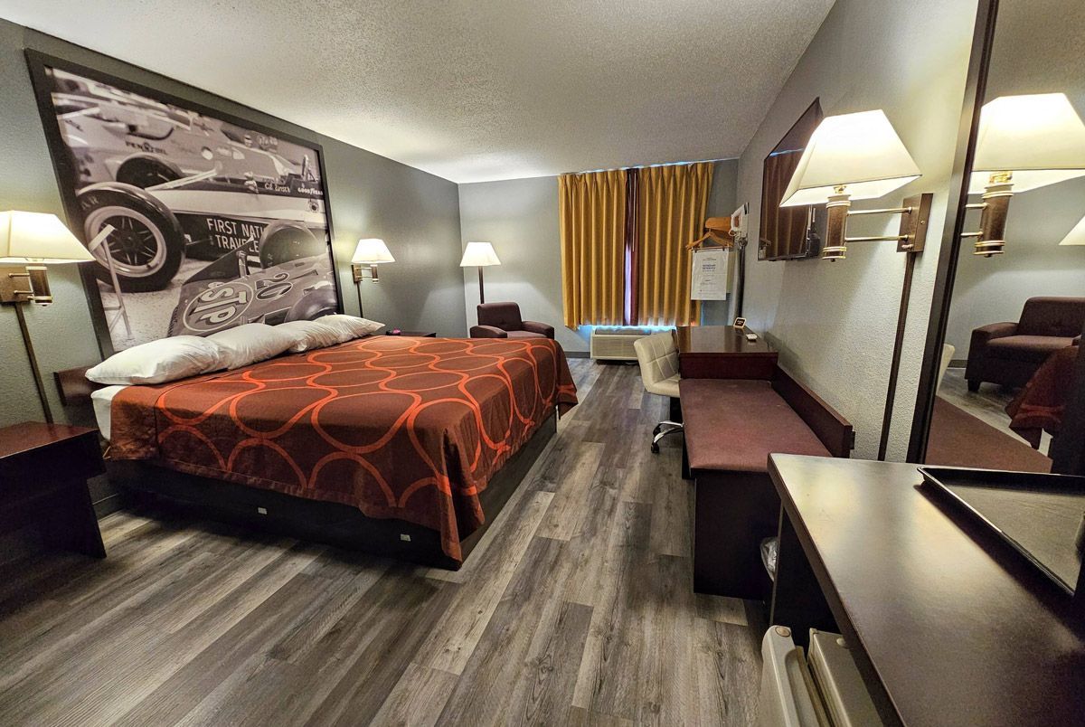 A hotel room with a large bed, brown patterned comforter, wood-look flooring, desk area, and wall-mounted art.