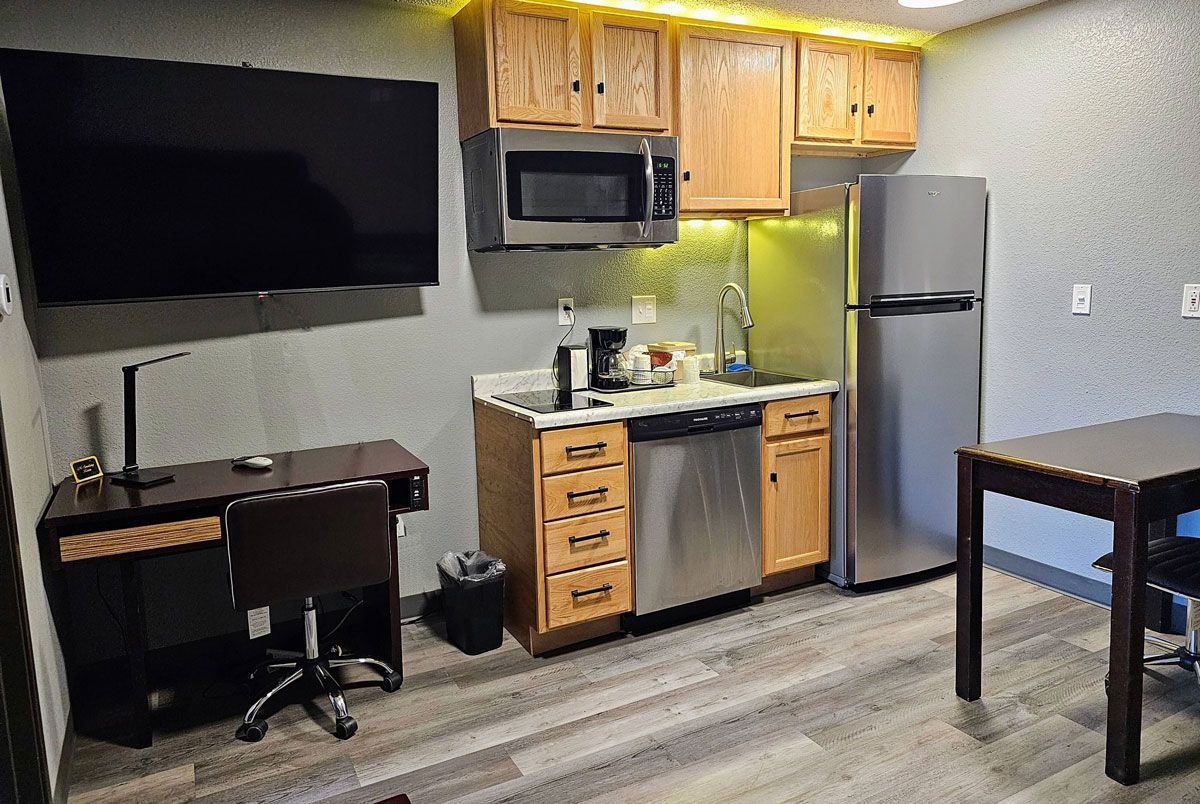 A hotel kitchenette with a desk, flat-screen TV, wooden cabinets, microwave, dishwasher, and a stainless steel fridge.