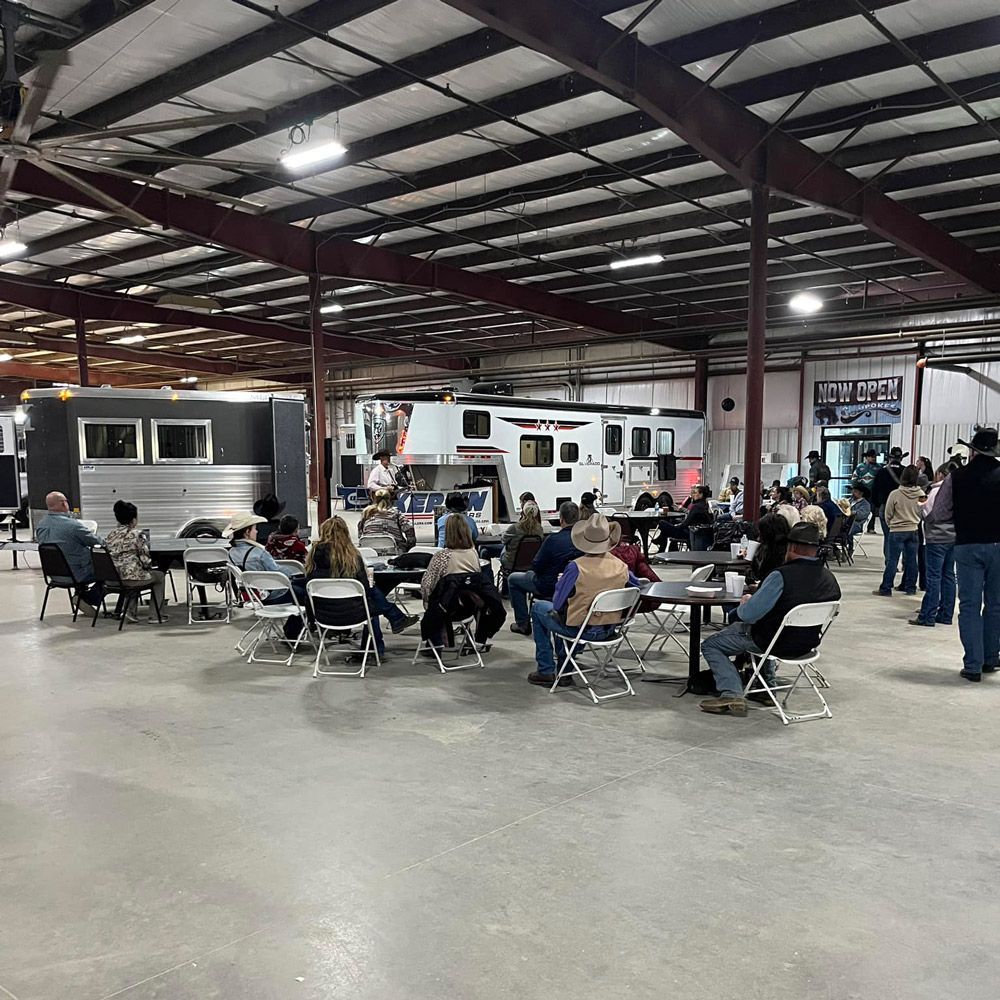 People sit at tables in a large indoor arena with trailers parked nearby, listening to a speaker during an event.