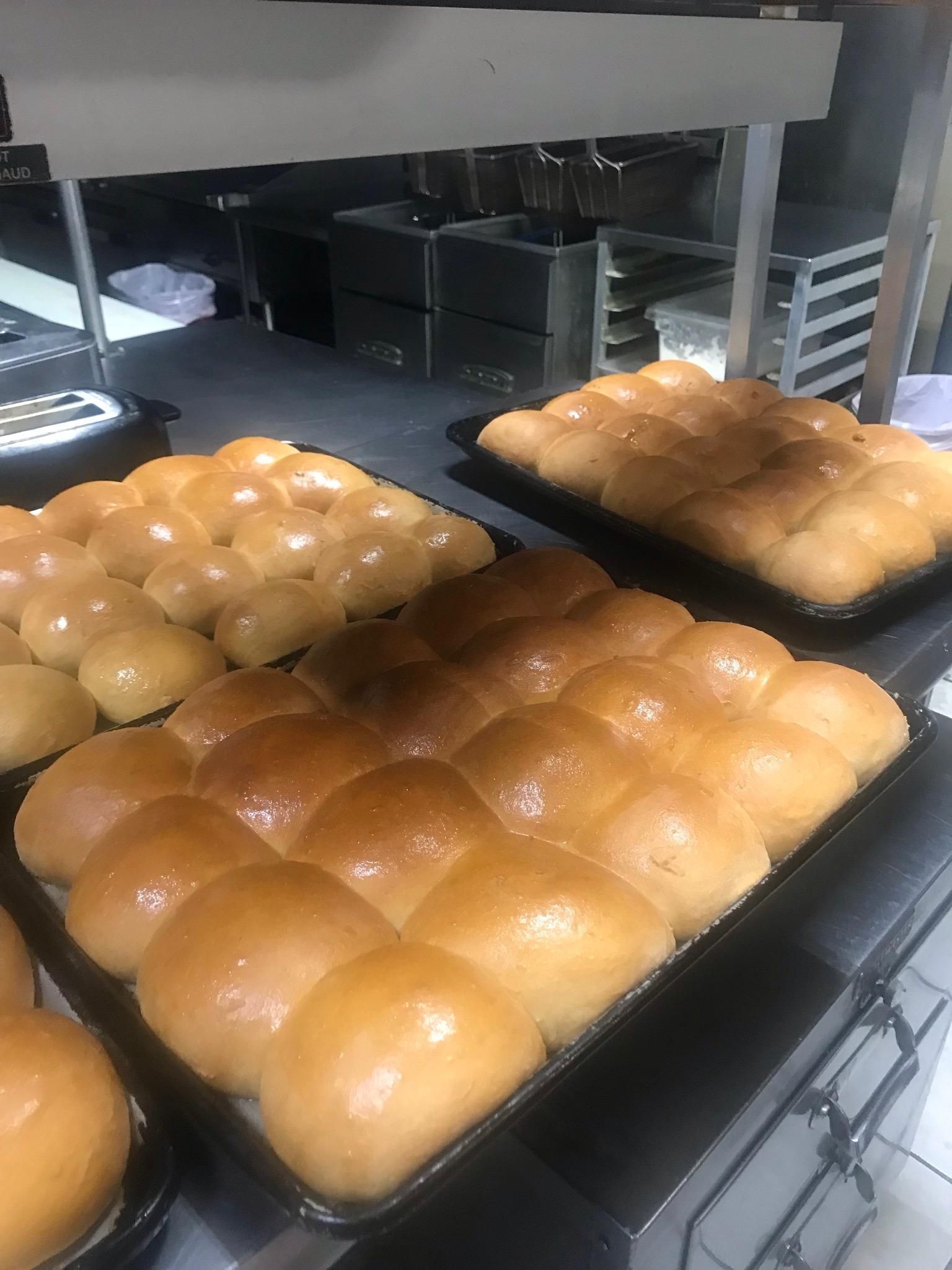 Several trays of freshly baked, golden-brown dinner rolls arranged in a commercial kitchen setting.