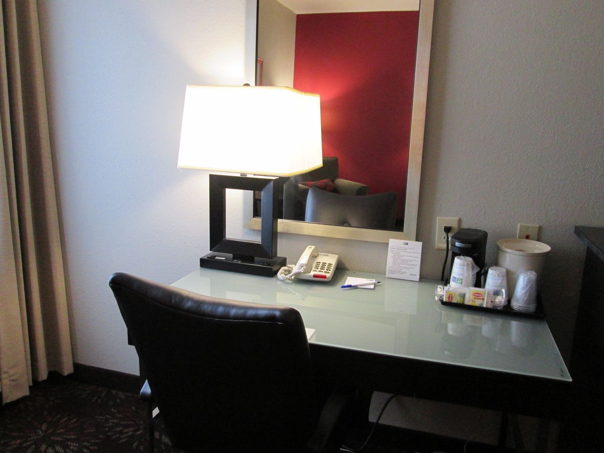 A hotel desk with a lamp, chair, mirror, and coffee service against a white wall.