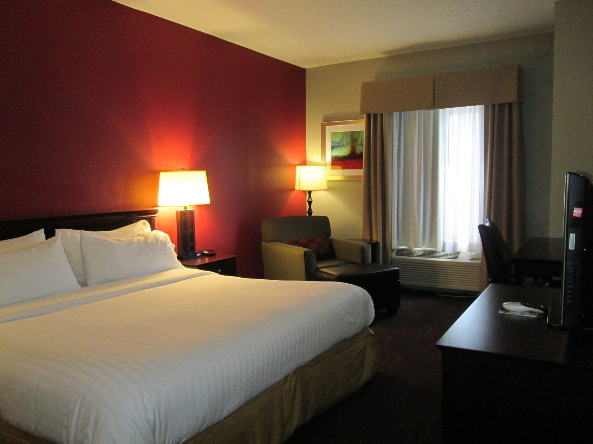 A hotel room featuring a large bed with white linens against a red wall, a side chair, window curtains, and a desk.