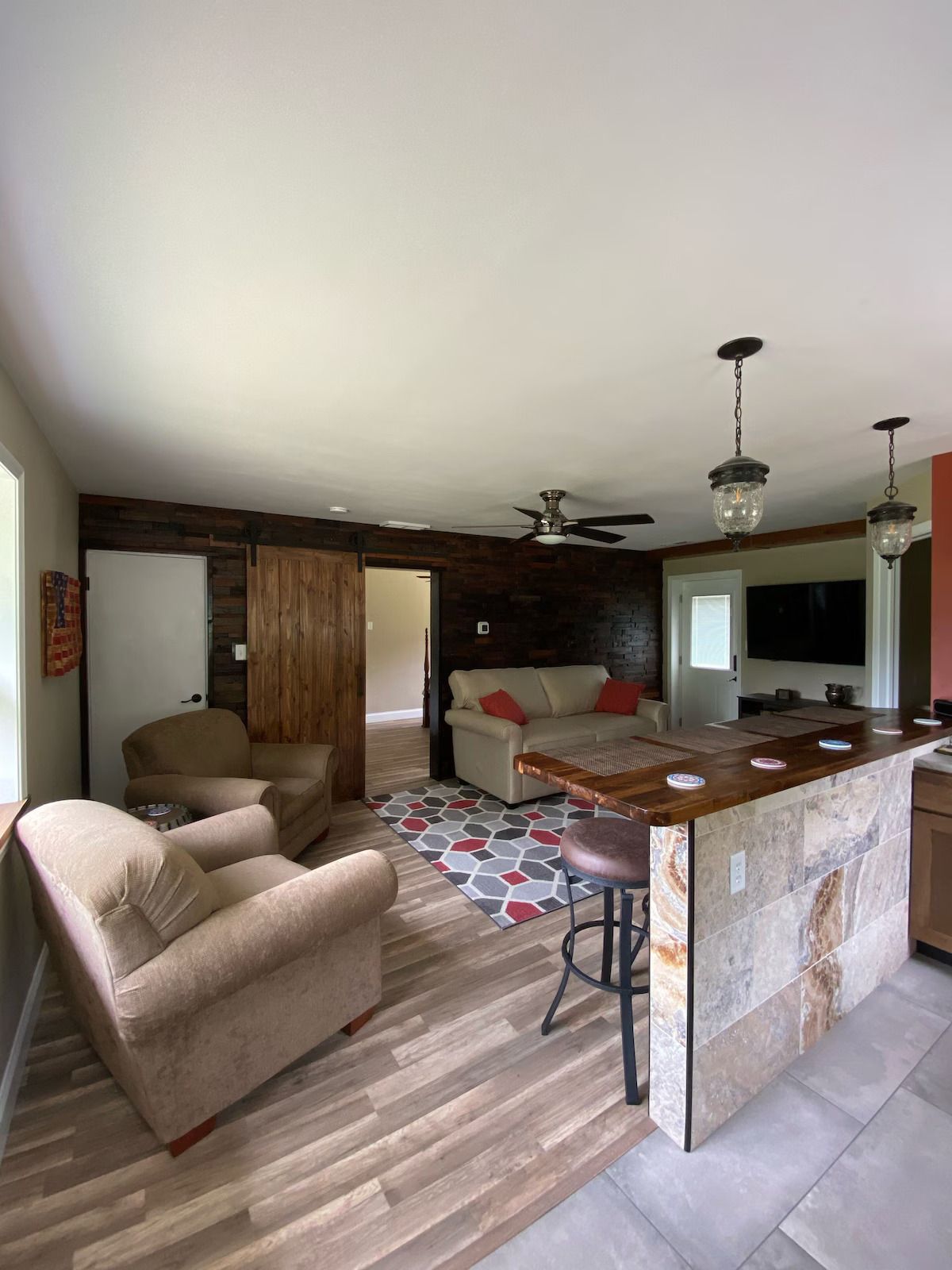 A living room and kitchen area featuring wood flooring, a stone-front bar, comfortable armchairs, and a sliding barn door.