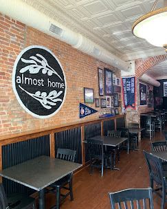 Cozy brick-walled cafe interior with black tables, framed wall art, and an “almost home” sign