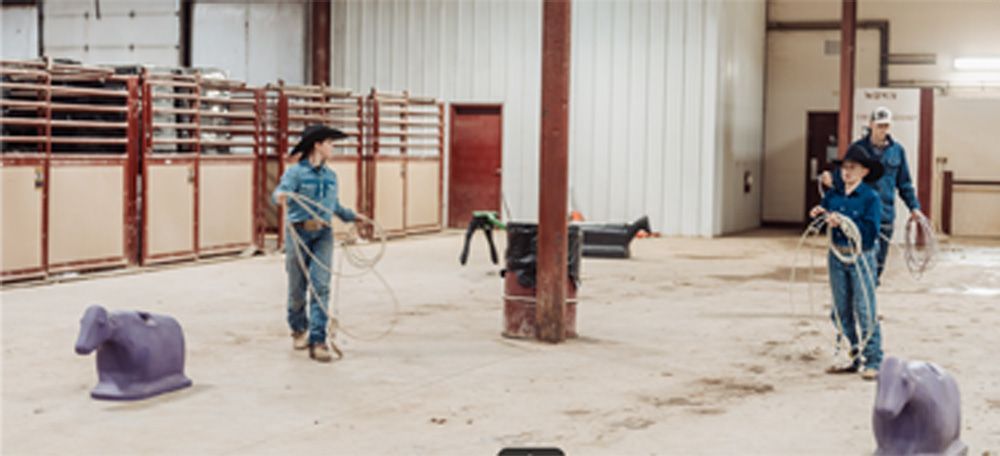 Two people roping in an indoor arena. Purple dummies and a wooden support beam are in the arena.