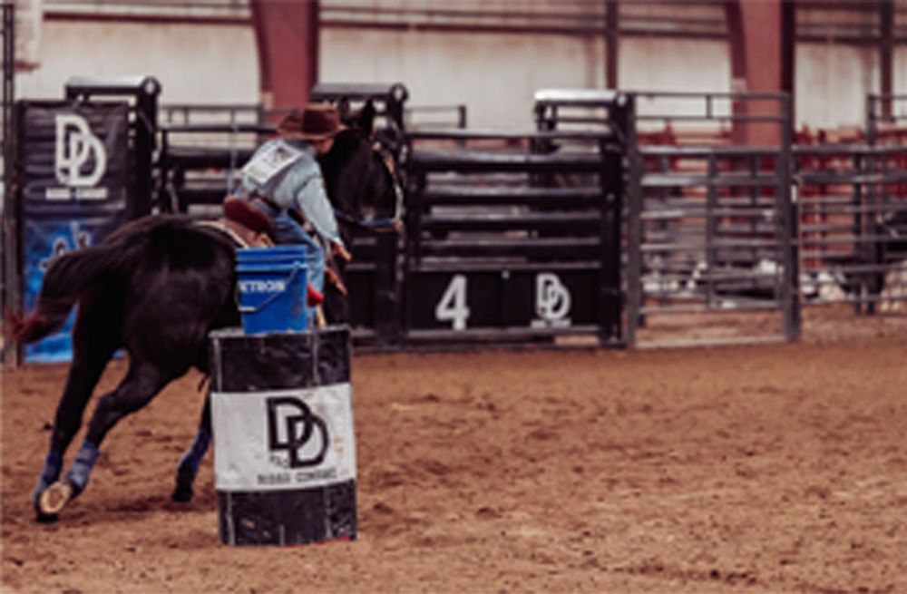 A person on a horse rounds a barrel in a rodeo arena. The barrel is black and white, and the arena is tan and brown.