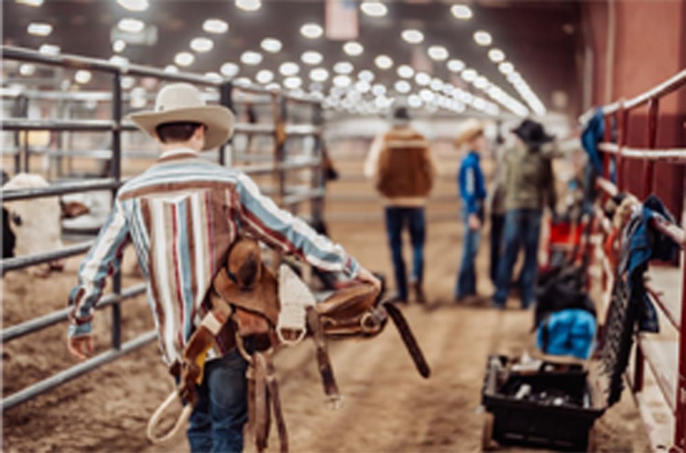 Boy in cowboy hat and shirt carrying a saddle in an arena; other people visible in the background.