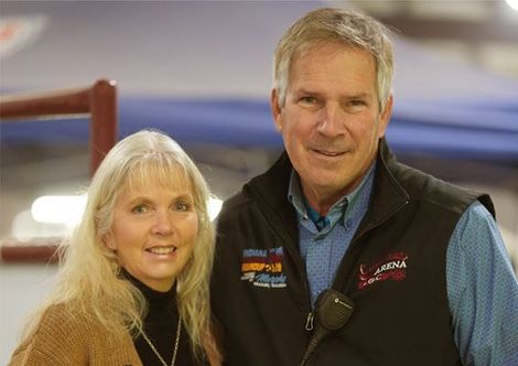 Couple smiling, posing together indoors. Woman with long blonde hair, man wearing a vest and blue shirt.