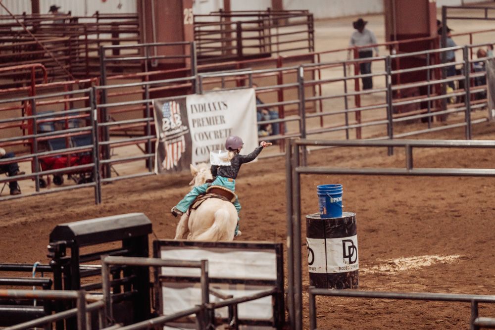 Cowgirl on a light horse rounds a barrel in a rodeo arena, raising her arm. Brown tones.