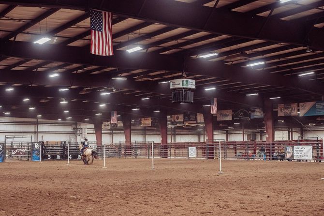 A rider on horseback moves through an indoor arena under bright lights, with an American flag hanging from the ceiling.