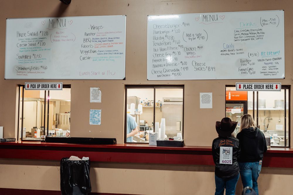 Two people at a food counter, ordering. Menu boards overhead. Kitchen workers visible through windows.