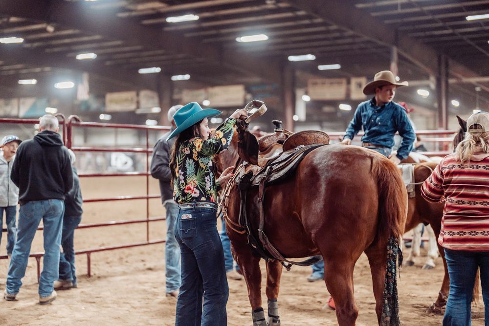 People preparing a horse for riding at an indoor rodeo; one person in a blue hat adjusts tack.