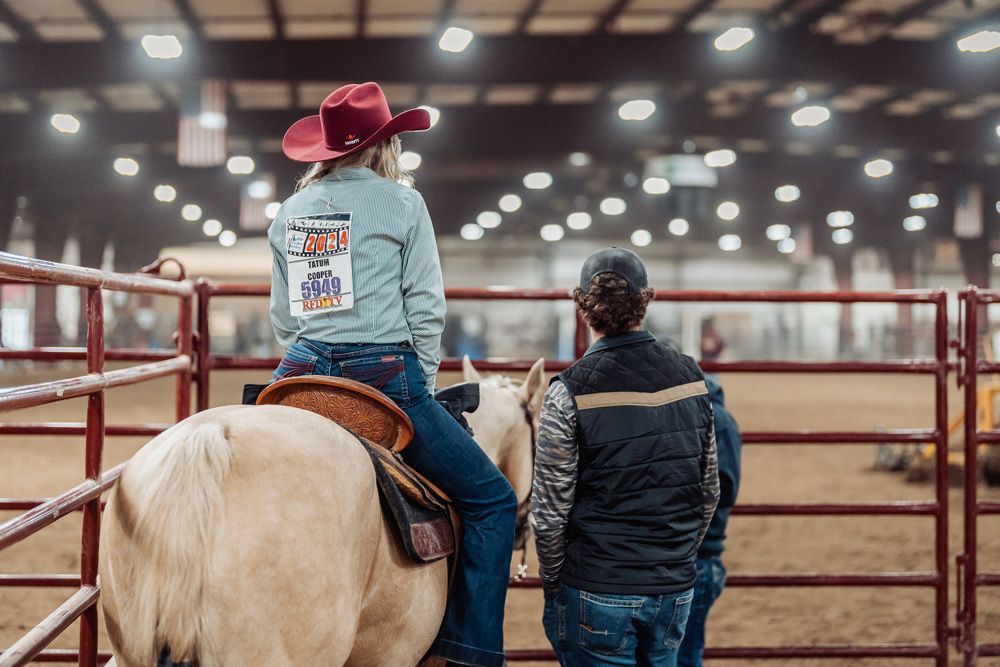 Woman on horseback in a rodeo arena, followed by another rider. Other riders and spectators in background.