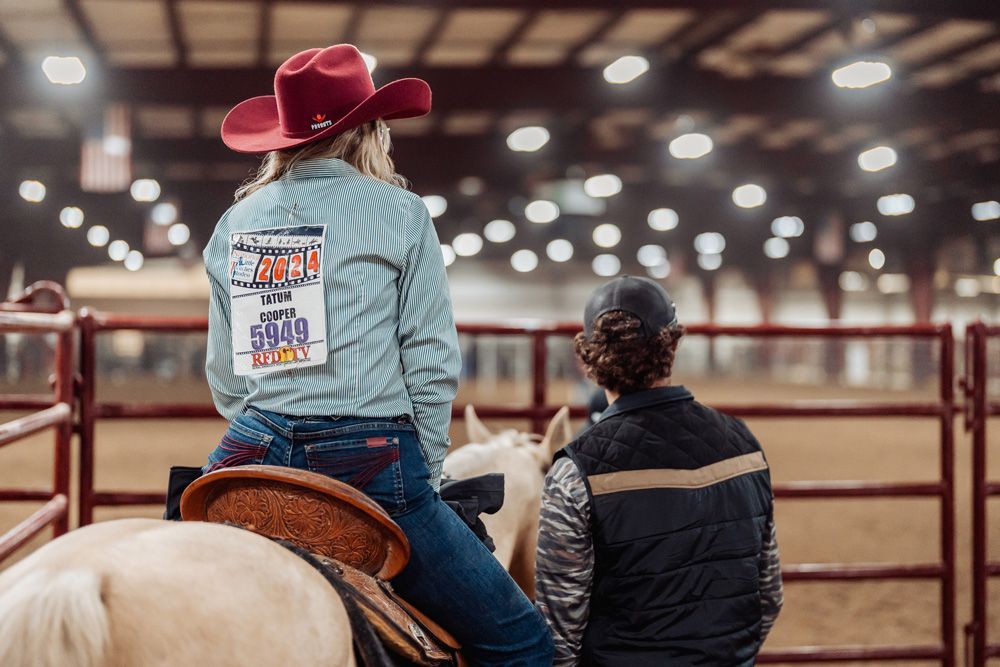 Woman on horseback wearing a red hat, looking at another person in a rodeo arena.