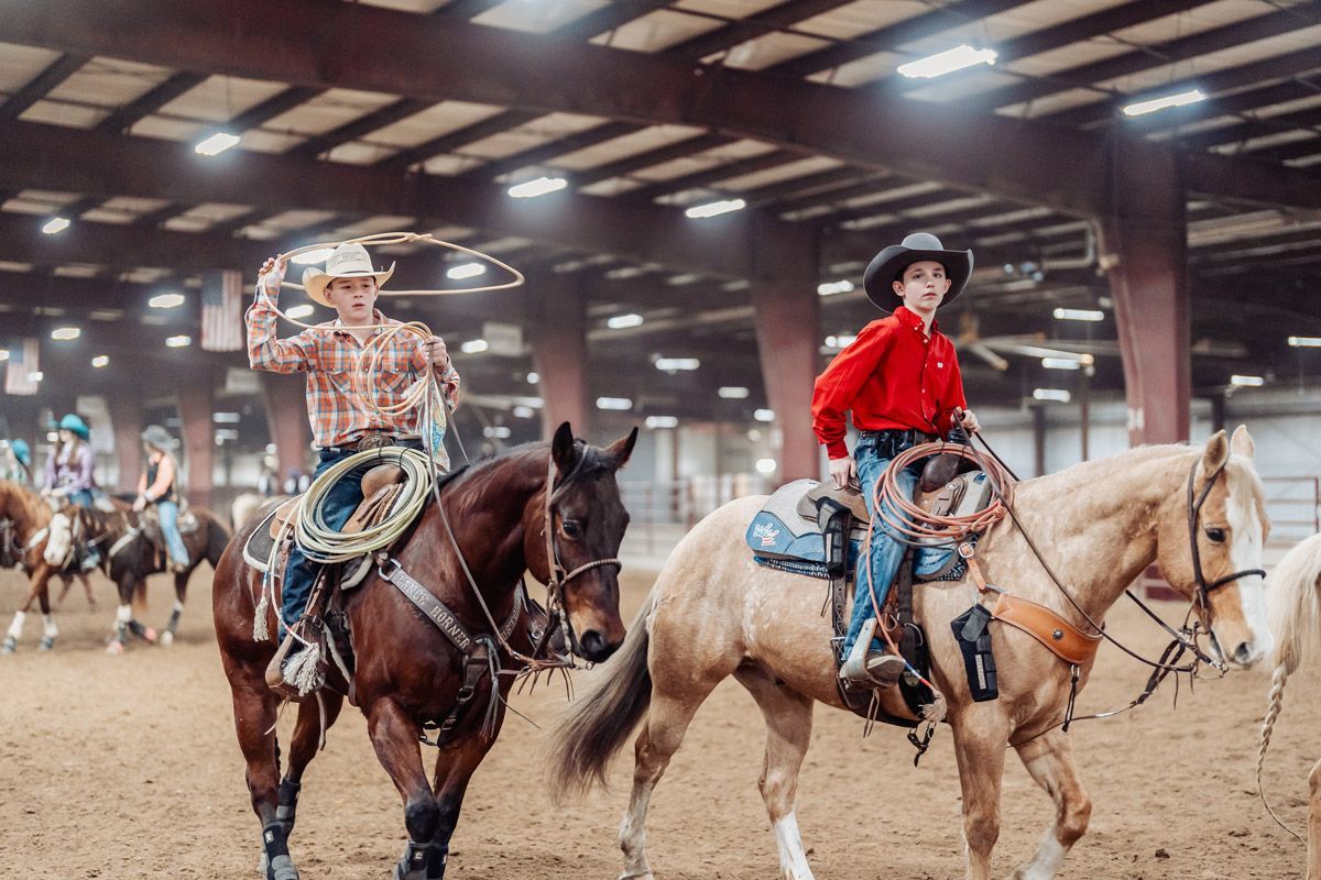 Two riders on horseback in an indoor arena, one twirling a lasso and the other riding a light-colored horse.