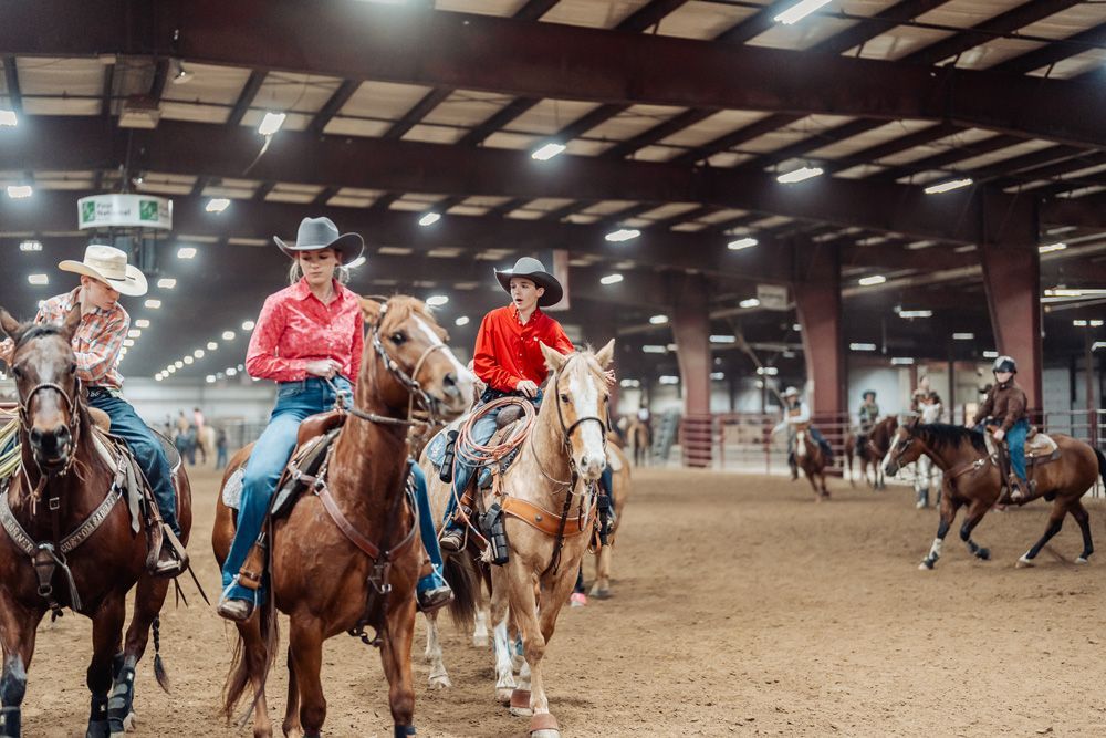 Cowgirls on horseback inside arena, wearing cowboy hats and western attire.