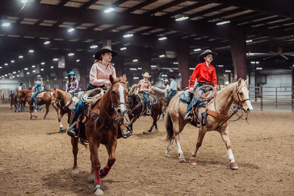 Cowgirls on horseback in an indoor arena, riding Western-style, preparing for competition.