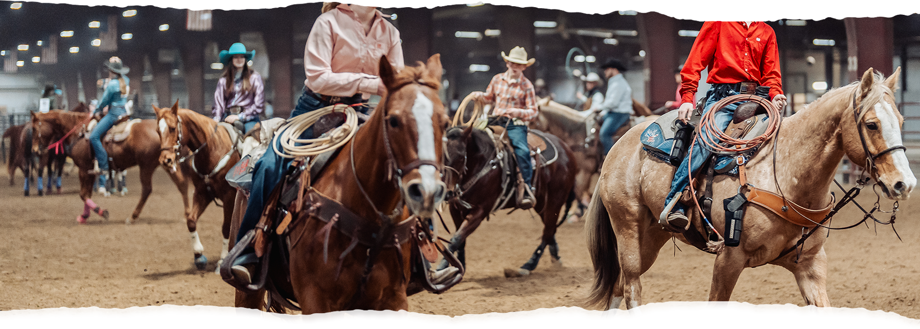 People riding horses in an indoor arena, wearing cowboy hats and western attire.