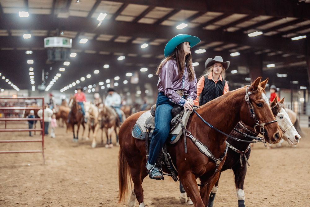 A person in a red shirt and cowboy hat riding a palomino horse in an indoor arena, other riders nearby.