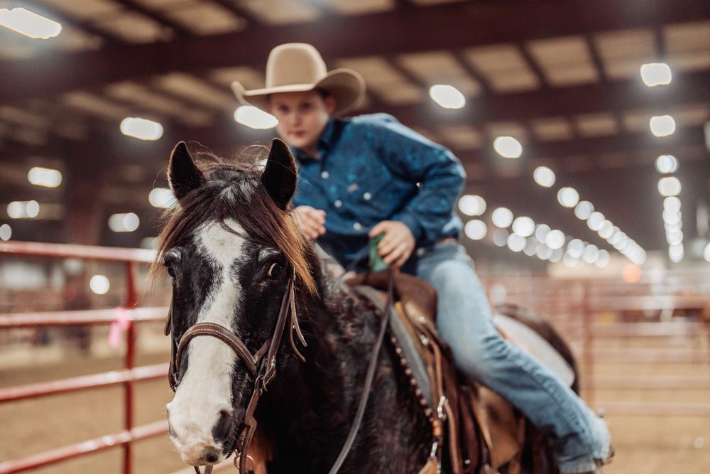 Young person in a cowboy hat and denim shirt with a horse in an indoor arena, looking at the camera.