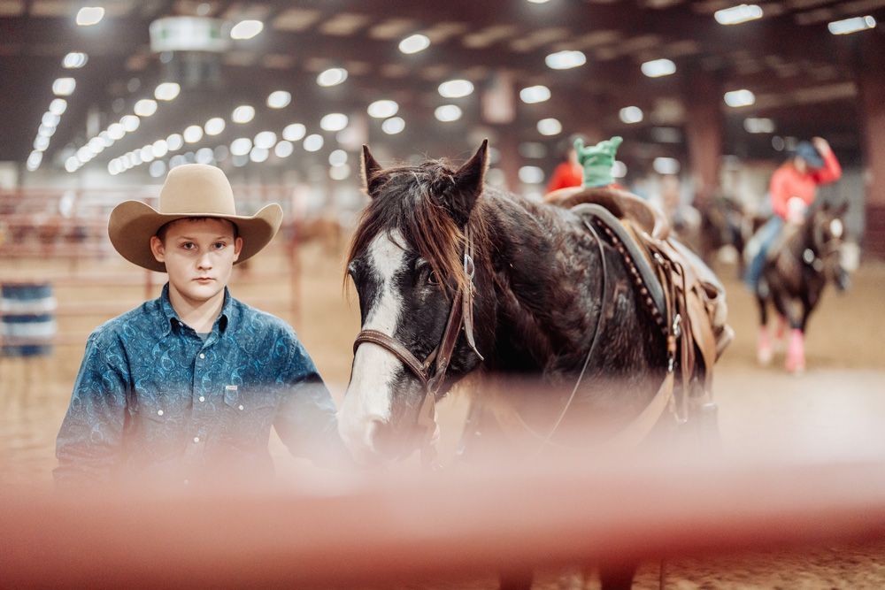 Person in cowboy hat stands with a saddled horse in an indoor arena. Other riders and horses are in the background.