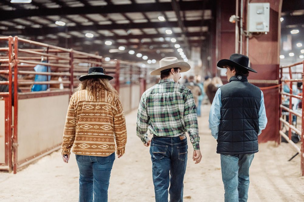 Three people in cowboy hats walk in a sandy indoor arena.
