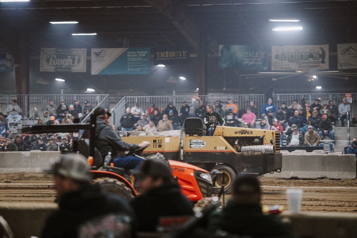 An orange tractor pulls a weighted sled in a dirt arena before a seated crowd under stadium lights.