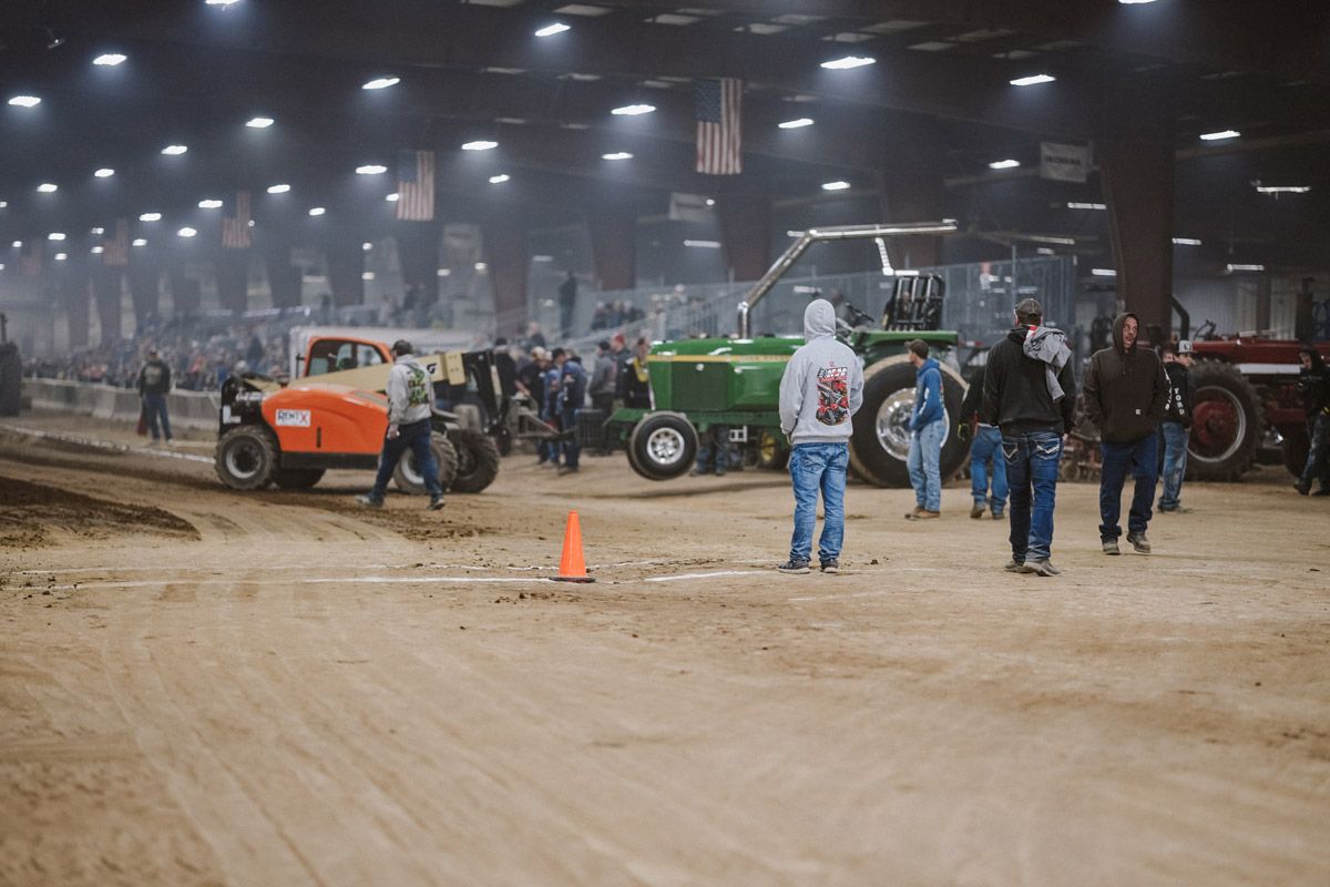 People gather in a large indoor arena around a green tractor and an orange tractor at a tractor pull event.