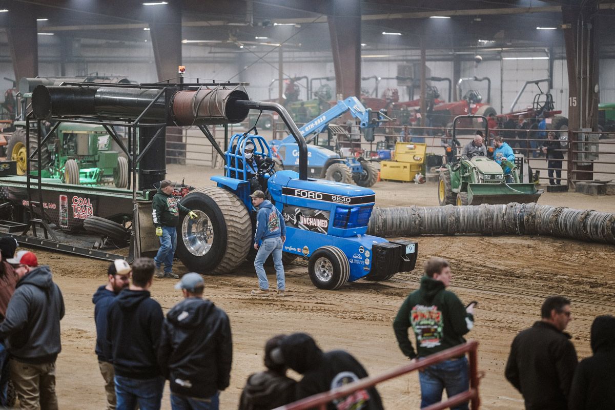 A bright blue tractor participates in an indoor tractor pull competition, with spectators standing in the foreground.