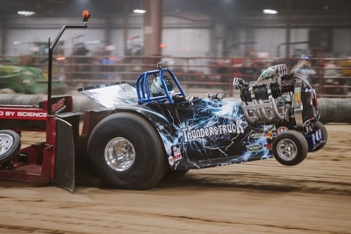 A modified tractor with a lightning graphic on its side pulls a weighted sled, lifting its front wheels off a dirt track.