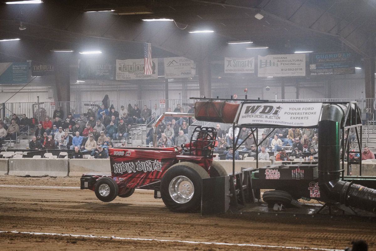 A red tractor pulling a weighted sled on a dirt track in an indoor arena filled with spectators.