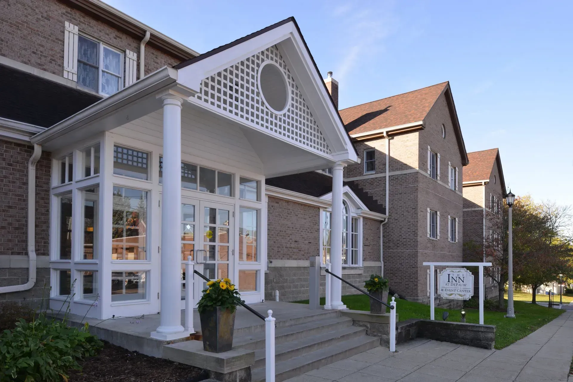 A brick building with a white portico entrance and stairs, set on a sunny day with green landscaping.