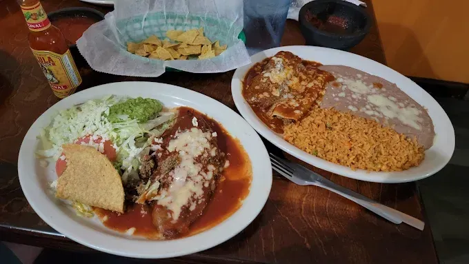 Plates of enchiladas and rice on a table, with a basket of chips and salsa in the background