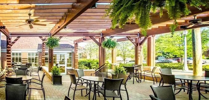 Covered outdoor patio with tables, chairs, hanging ferns, and sunlight streaming through wooden beams.