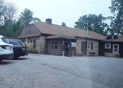 Stone building with brown roof and parked cars in a gravel lot, under cloudy skies