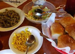Assorted dishes on a table: bread rolls, corn, green beans, and a salad bowl with serving spoons.