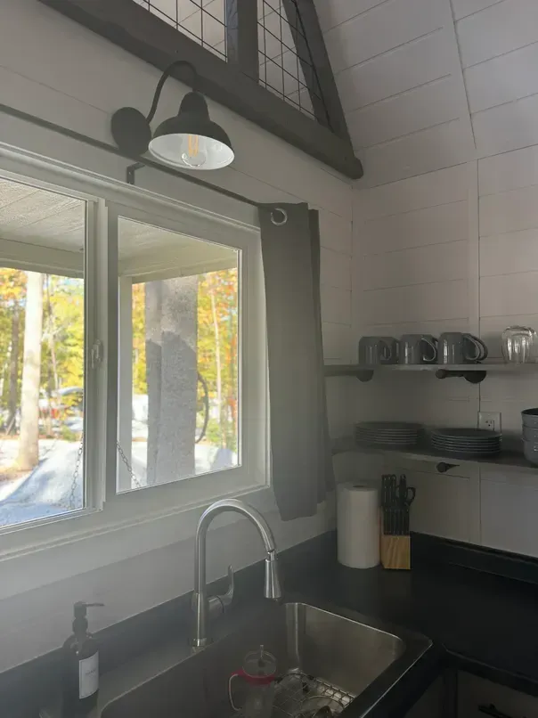 A kitchen sink and faucet under a window, with open shelving, mugs, and a paper towel holder on a white-tiled wall.
