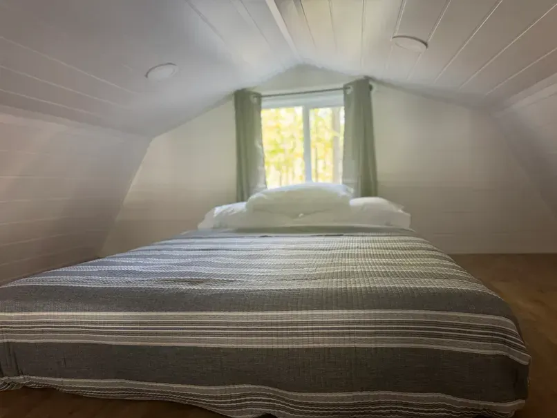 A mattress with a striped gray and white blanket and white pillows inside a small attic room with sloped white walls.