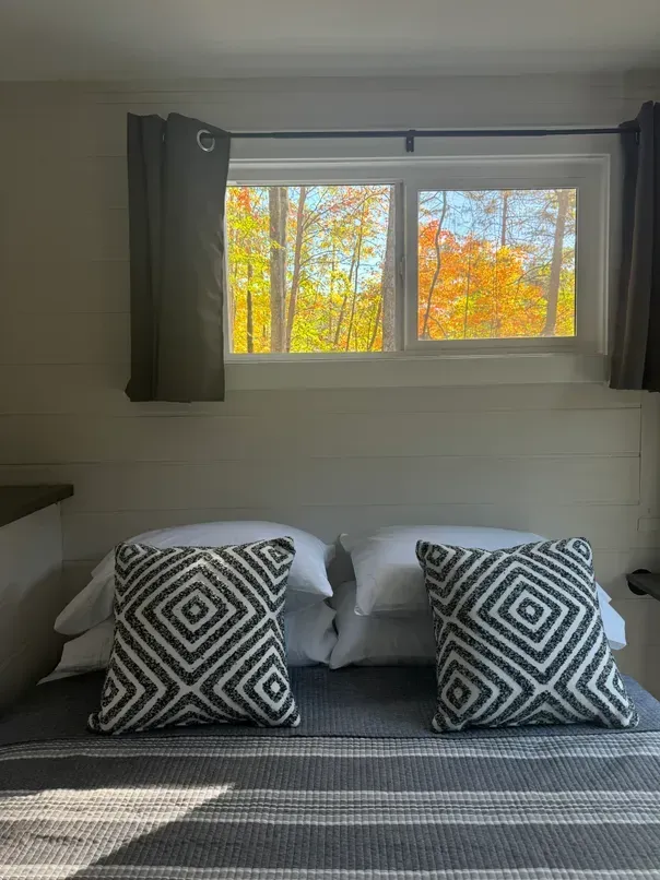 A bed with grey bedding and two geometric pillows sits beneath a window showing an autumn forest.
