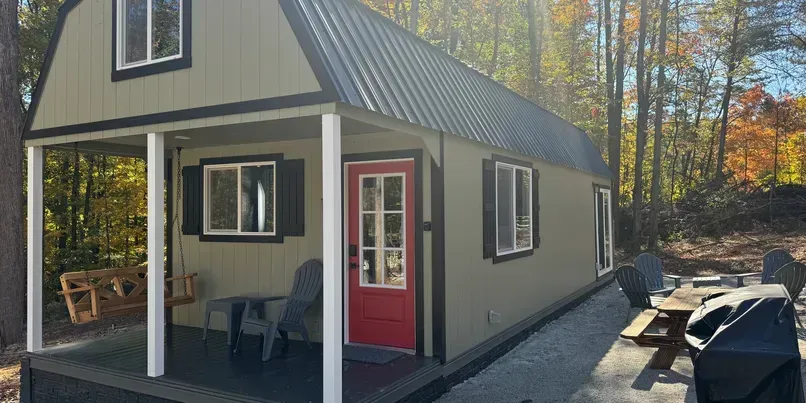 A beige tiny house with a metal roof, red door, and front porch in a wooded setting, with an outdoor table nearby.