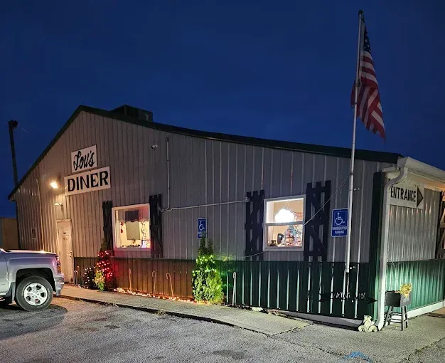 An exterior view of a silver metal diner at dusk, featuring a sign, an American flag, and an entrance with ramp access.