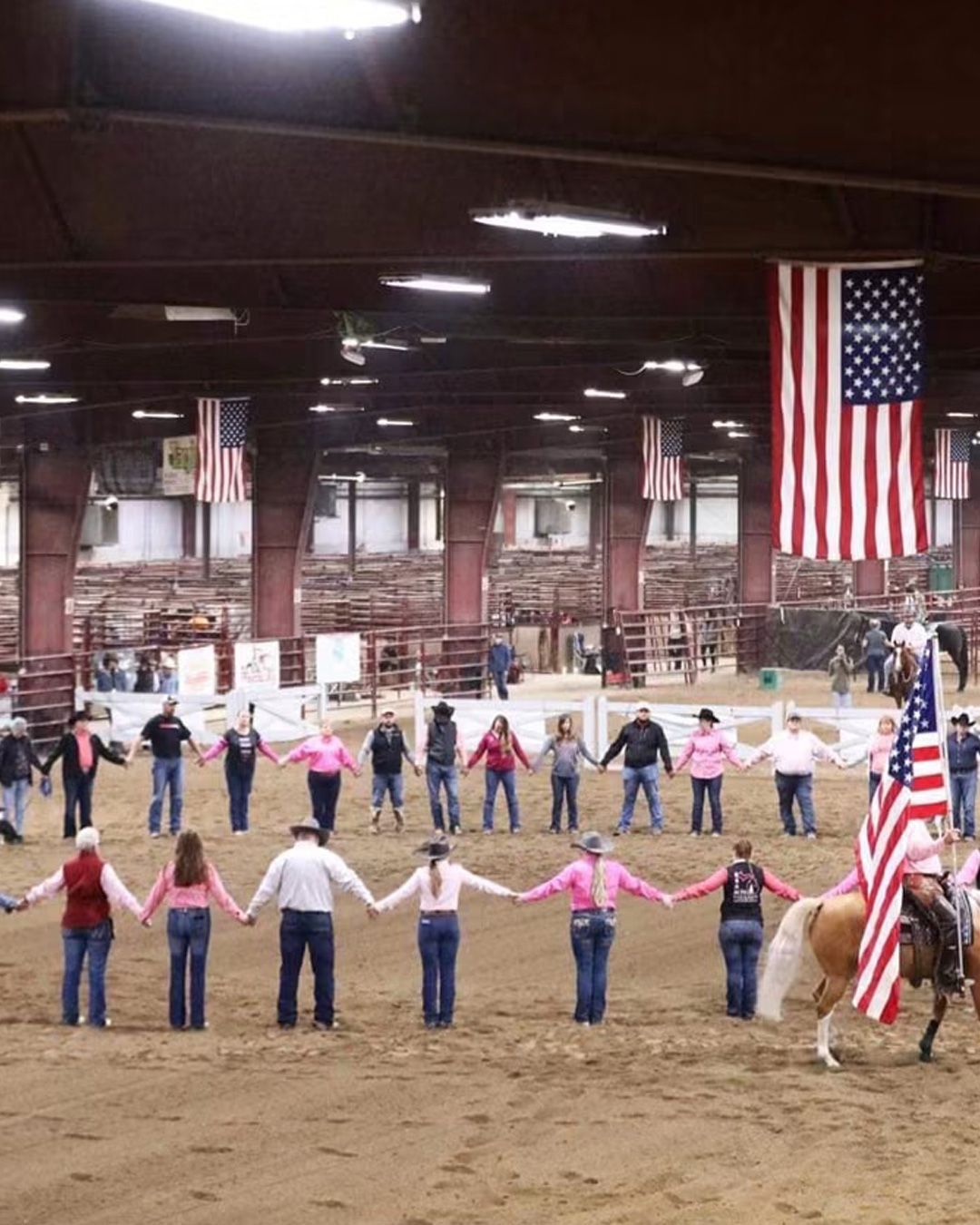 People holding hands in a circle inside an arena, with large American flags hanging overhead.