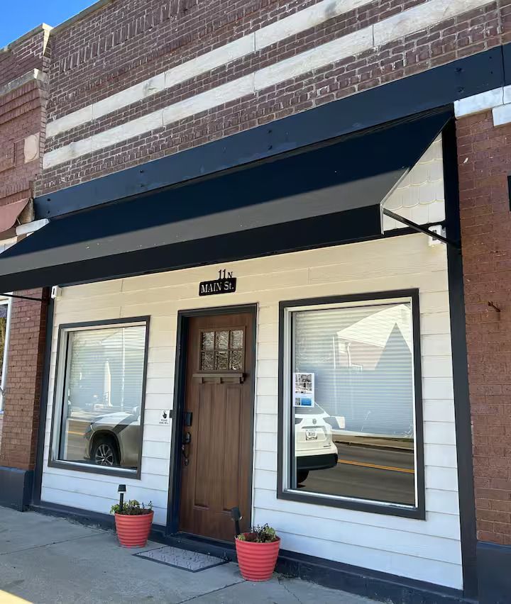 A storefront with white horizontal siding, a dark brown wooden door, large windows, and a black awning on a brick building.